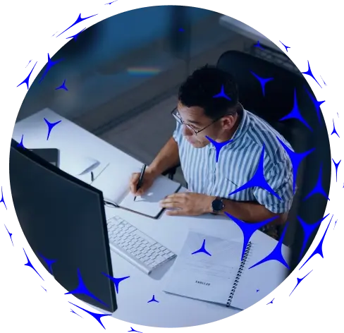 Pessoa trabalhando em mesa branca, escrevendo em caderno ao lado de computador e teclado, vista de cima, com elementos gráficos azuis ao redor.