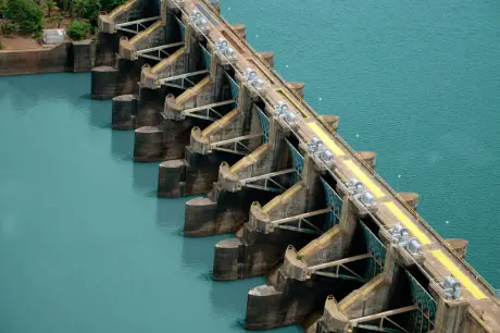 Vista aérea de uma barragem com comportas alinhadas em estrutura de concreto, em um reservatório de tom azul-esverdeado.