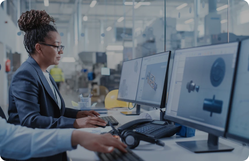Pessoa trabalhando em uma estação com dois monitores, analisando modelos e gráficos digitais em um ambiente tecnológico ou industrial.