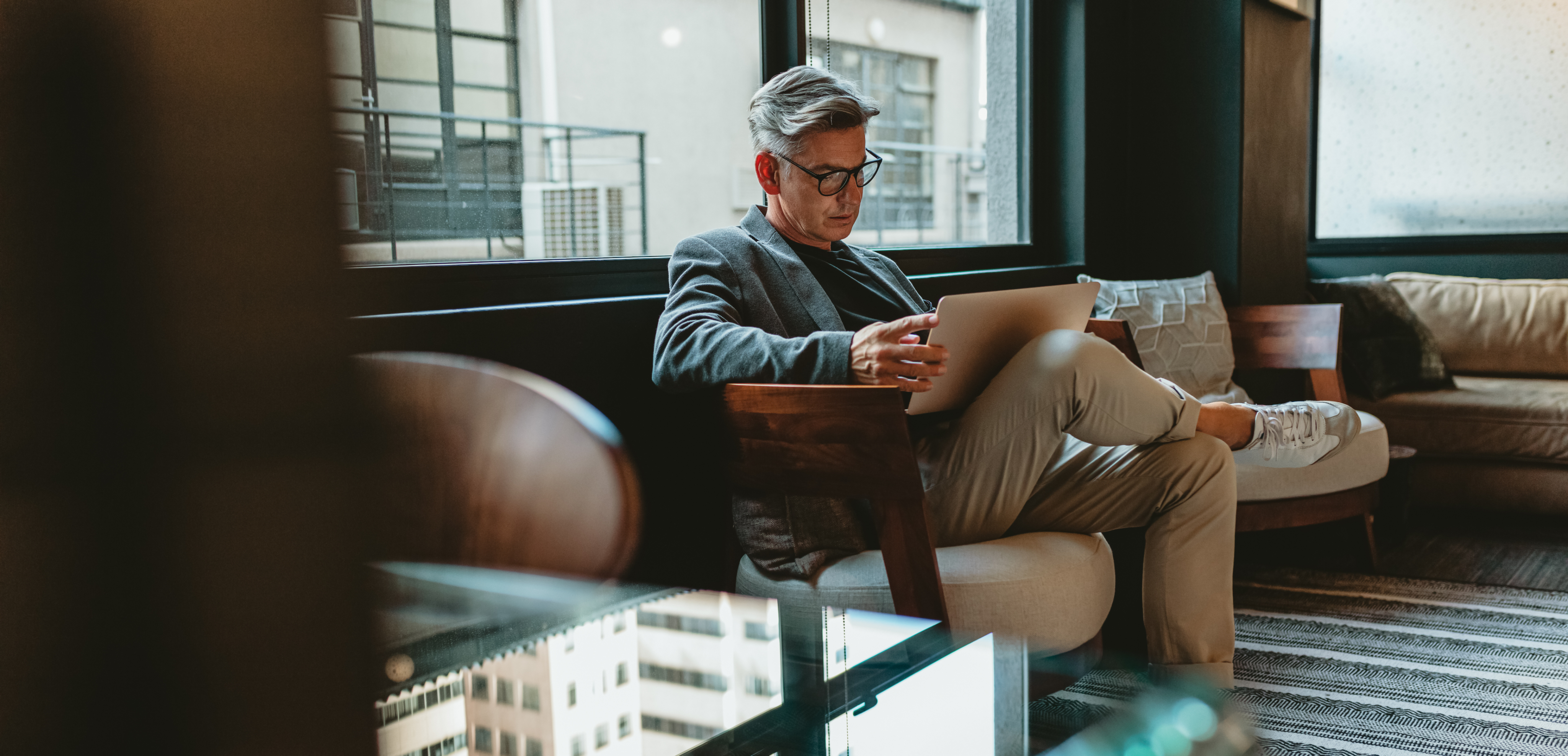 Man with glasses sitting on a modern armchair in a corporate setting, working on a laptop.