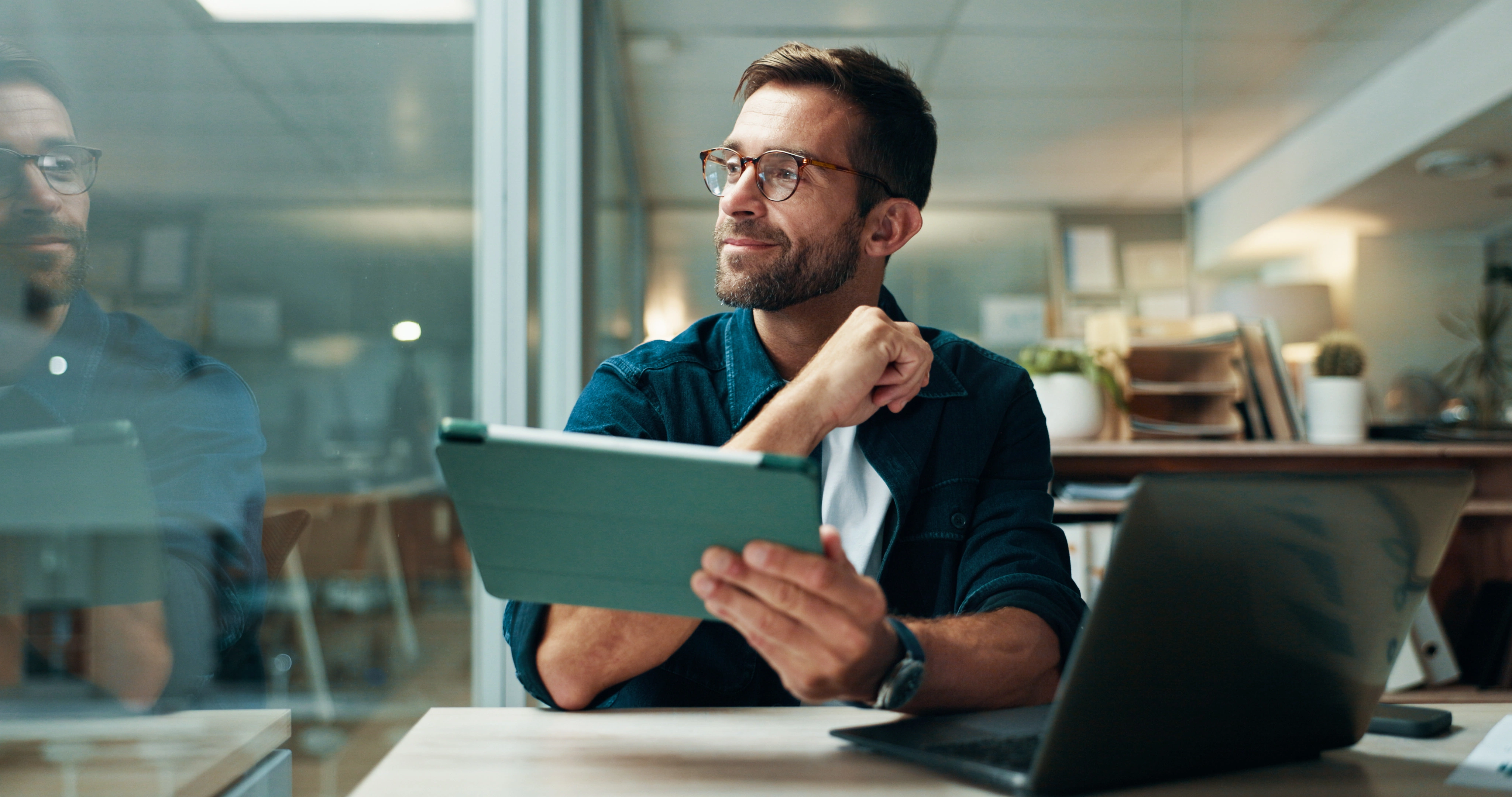 Man with glasses sitting in an office, holding a tablet and looking thoughtfully to the side, with an open laptop in front of him.