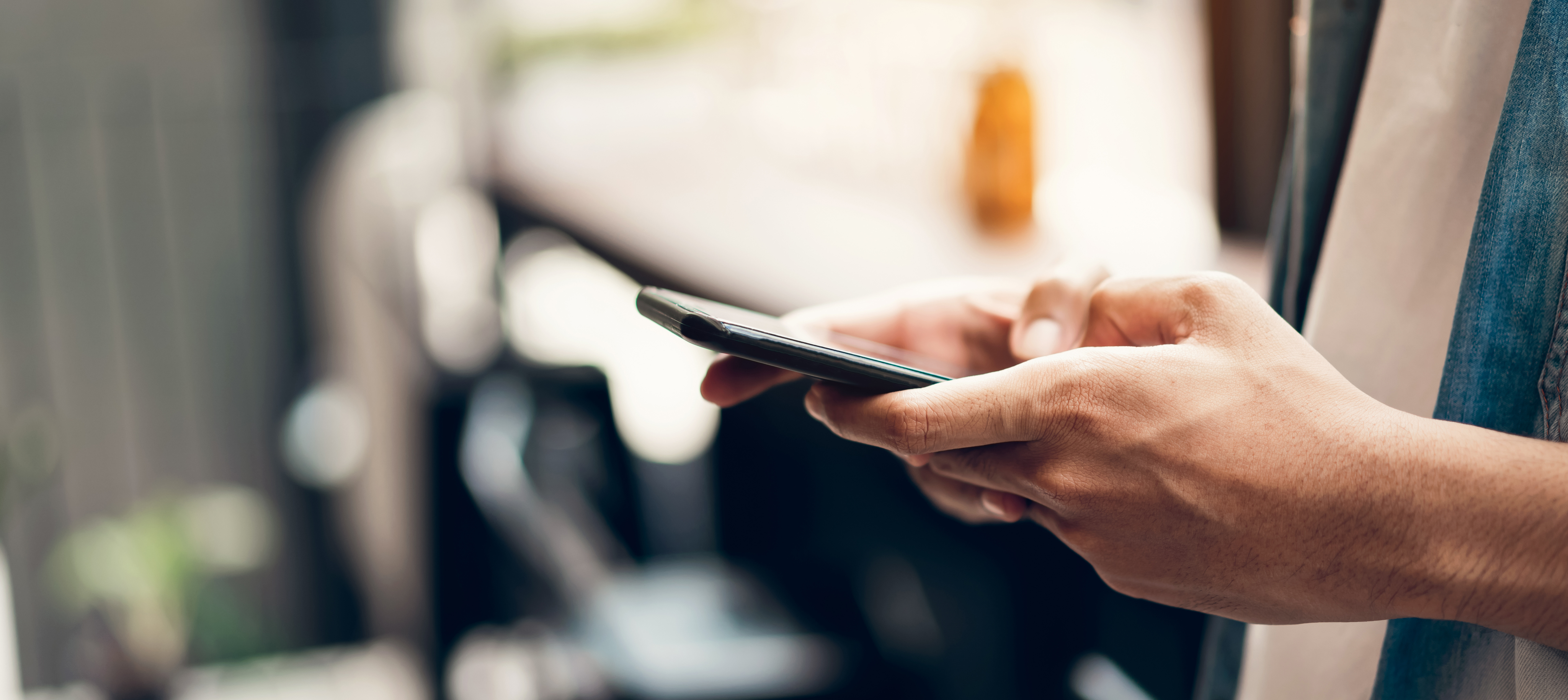 Close-up of a person's hands holding a smartphone, while using their thumb to tap and navigate the screen, against a blurred background with soft, warm lighting.