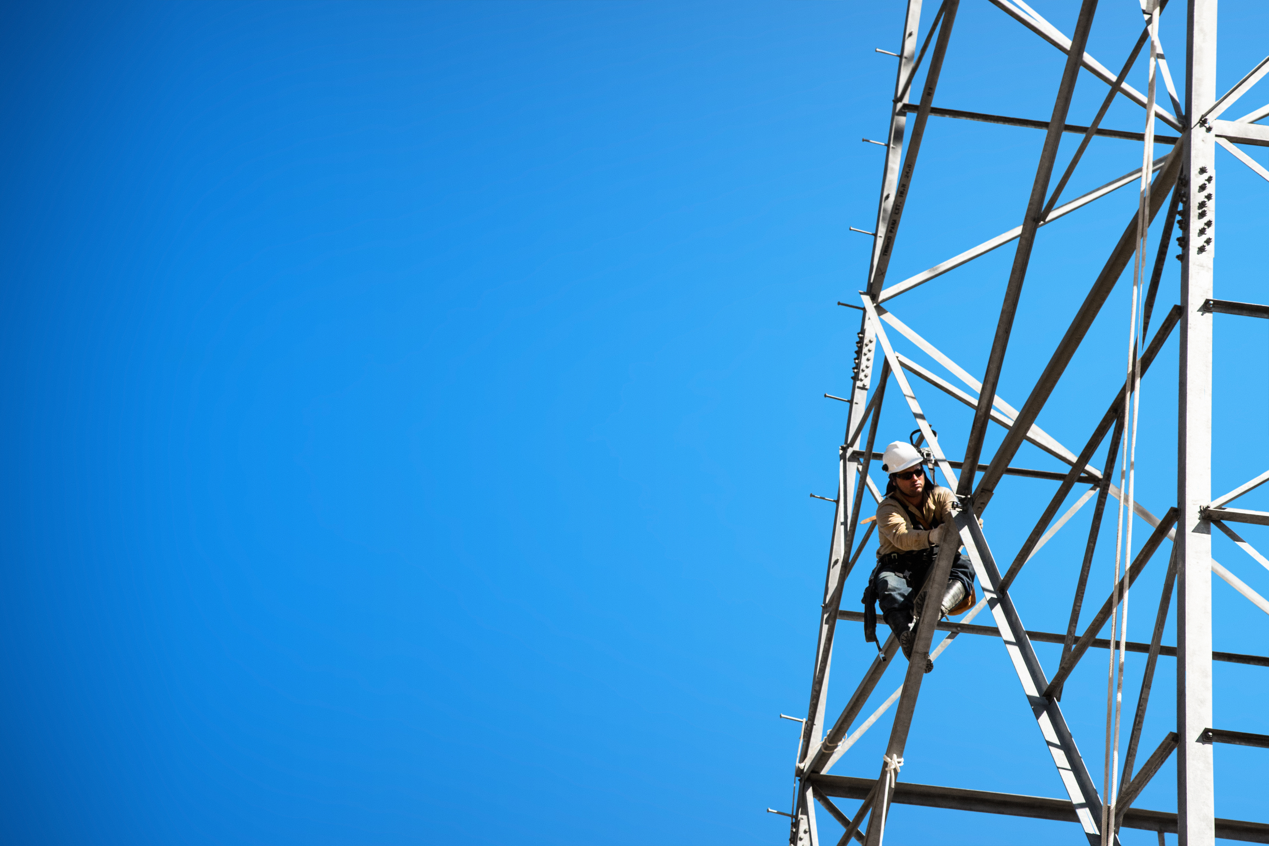Worker with safety equipment climbs a metal tower against a clear blue sky.