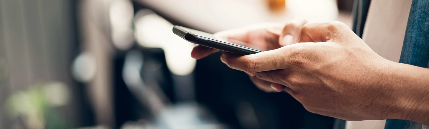 Close-up das mãos de uma pessoa segurando um smartphone, enquanto usa o polegar para tocar e navegar na tela, com um fundo desfocado e iluminação suave e quente.
