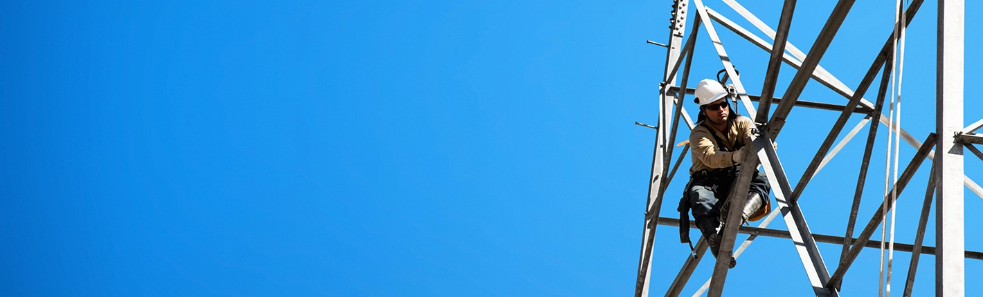 A worker wearing safety equipment climbs a metal tower against a clear blue sky.