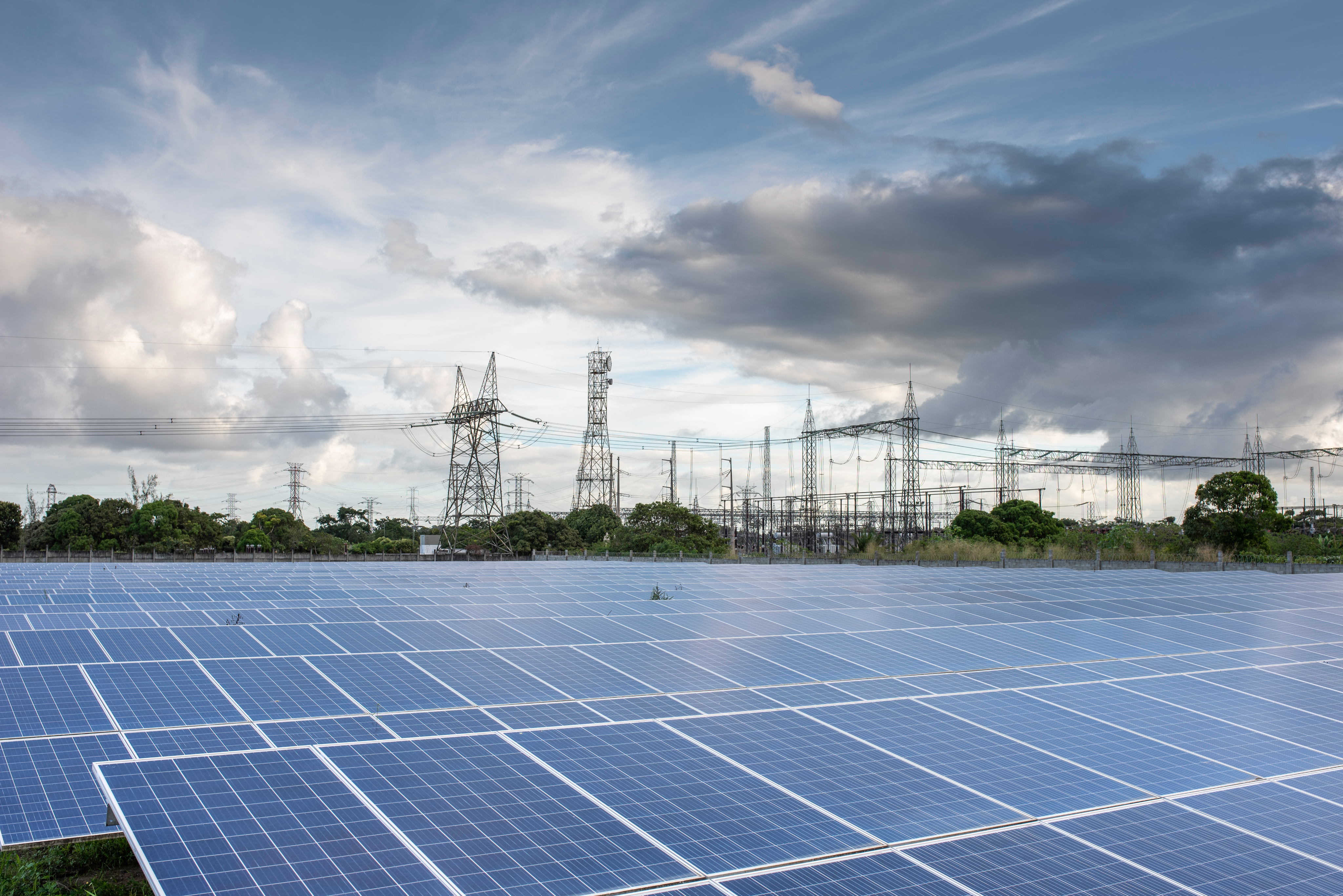 A large field of solar panels at a photovoltaic power plant in the foreground, with a substation and power transmission towers in the background, under a dramatic cloudy sky.