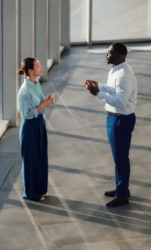 Duas pessoas em um ambiente corporativo conversam em pé próximo a grandes janelas, em espaço interno com iluminação natural.