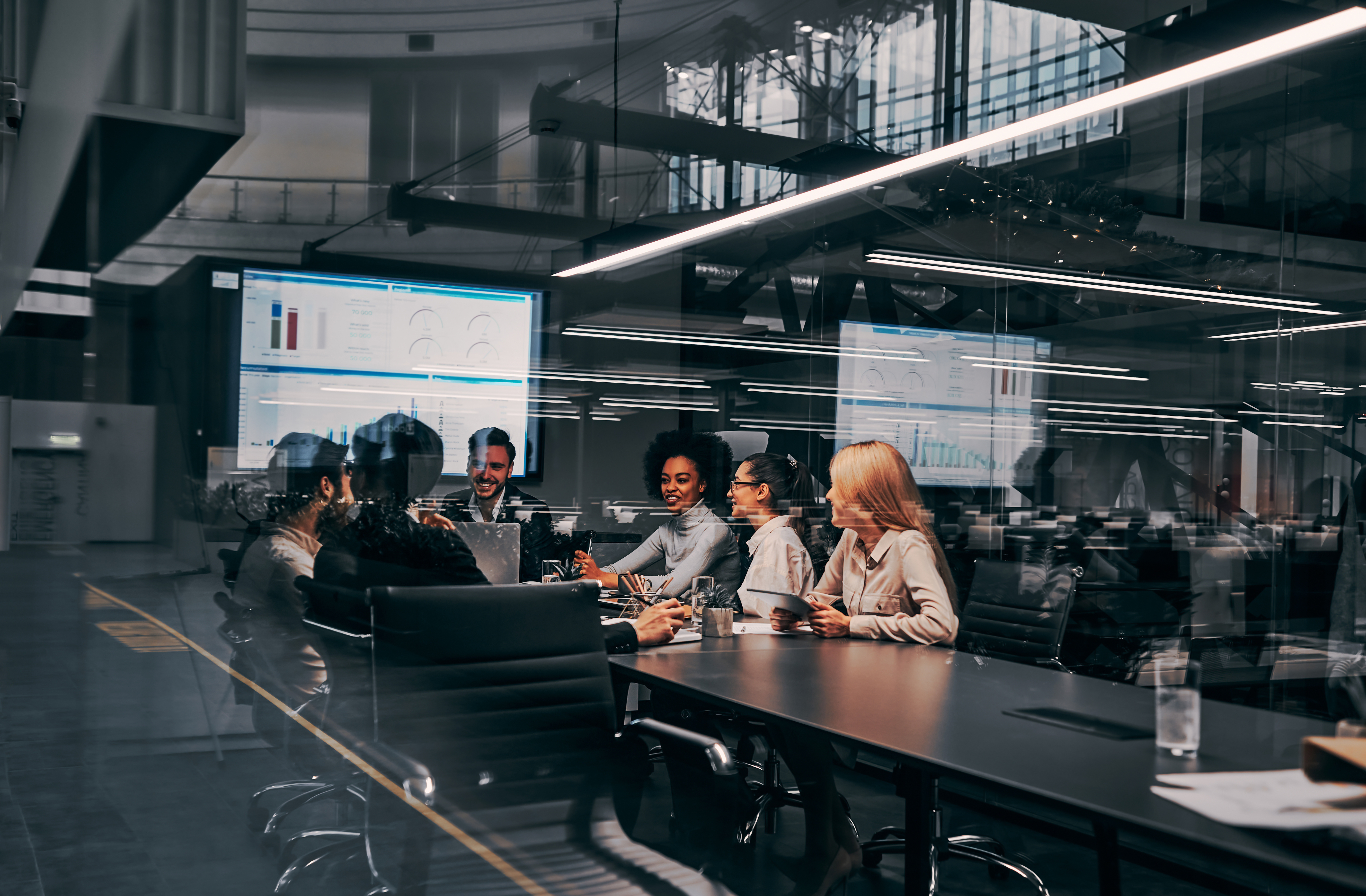 Team of professionals in a corporate meeting room with screens displaying charts and reports.