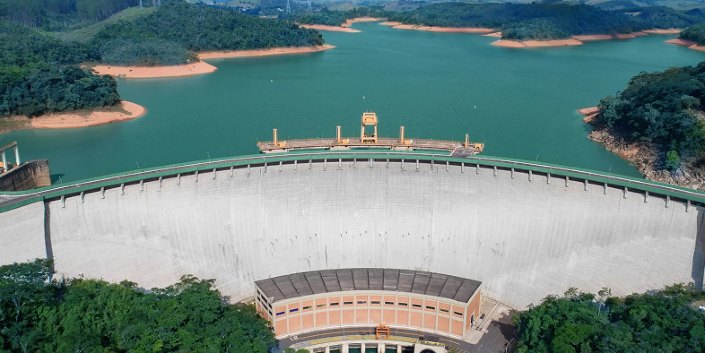 ista aérea de uma grande barragem de concreto em arco, retendo um reservatório de água esverdeada cercado por colinas arborizadas.