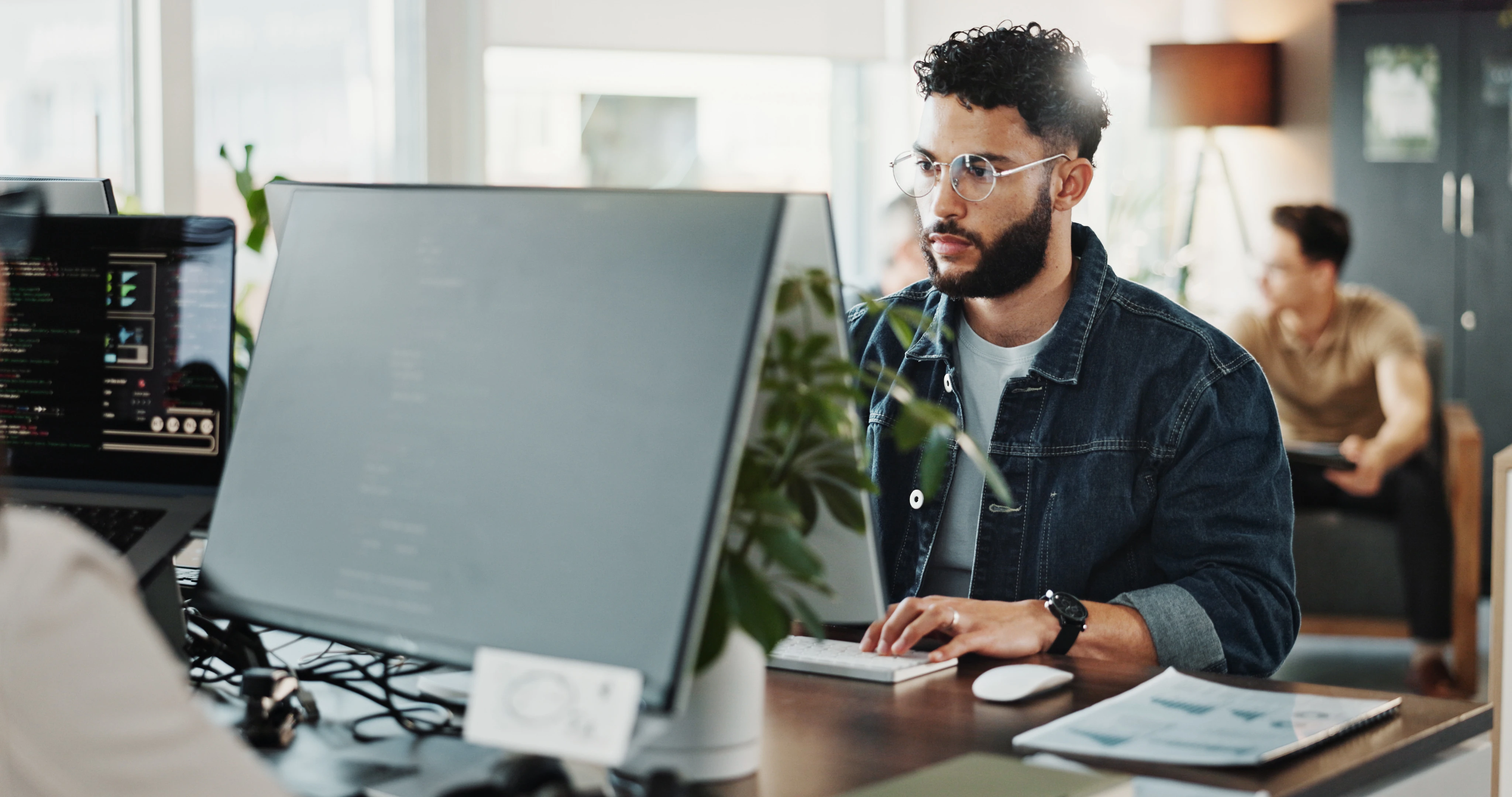Homem pardo de cabelos cacheados e óculos trabalha olhando para uma tela de computador
