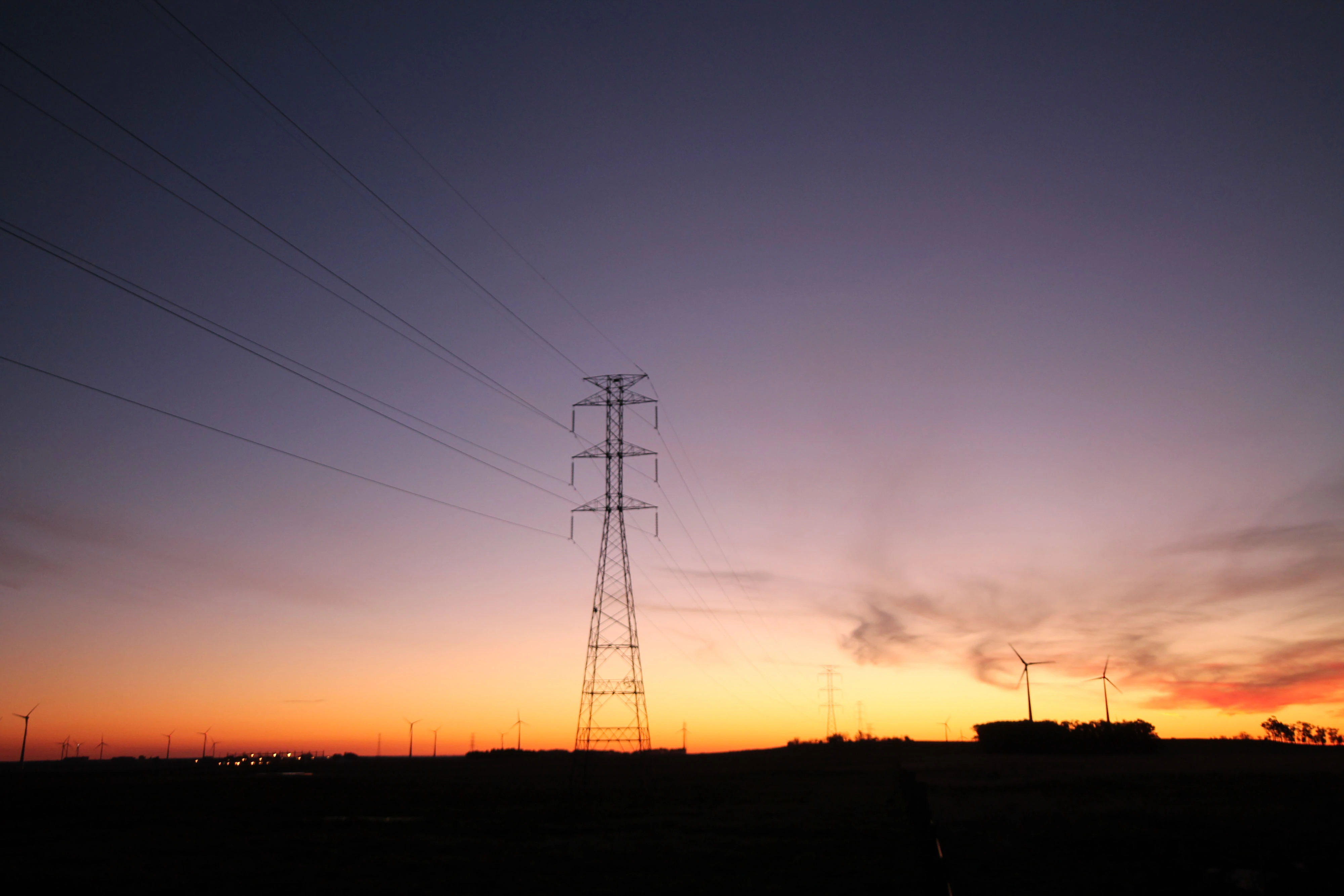 Landscape at dusk with the silhouette of a power transmission tower in the center and several wind turbines in the background. The sky displays a color gradient from orange to purple.