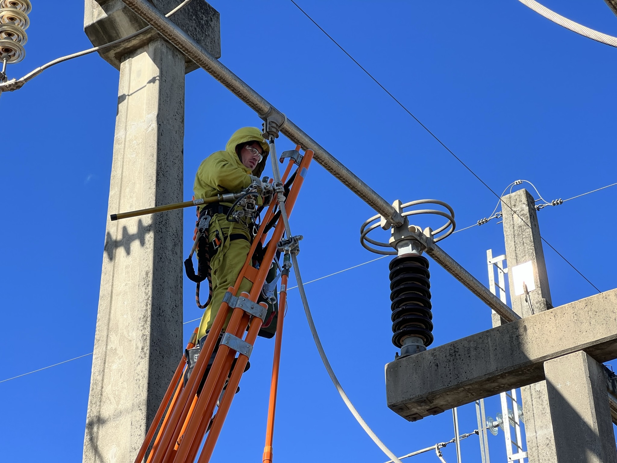 High-voltage electrician, wearing a full yellow protective suit and helmet, positioned on an orange ladder while performing maintenance on a busbar in a power substation, with a blue sky in the background.