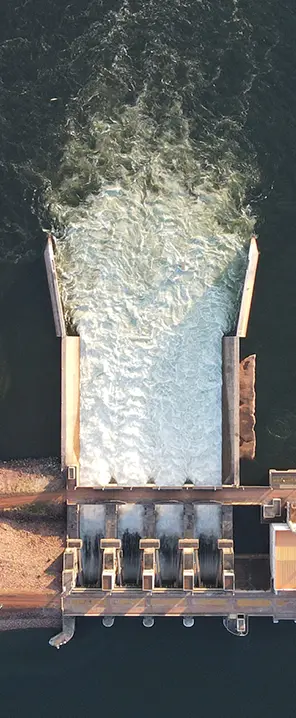 Imagem aérea de uma usina hidrelétrica, mostrando a água sendo liberada pelo vertedouro ao centro, com estruturas de concreto, linhas de transmissão e áreas de terra ao redor.