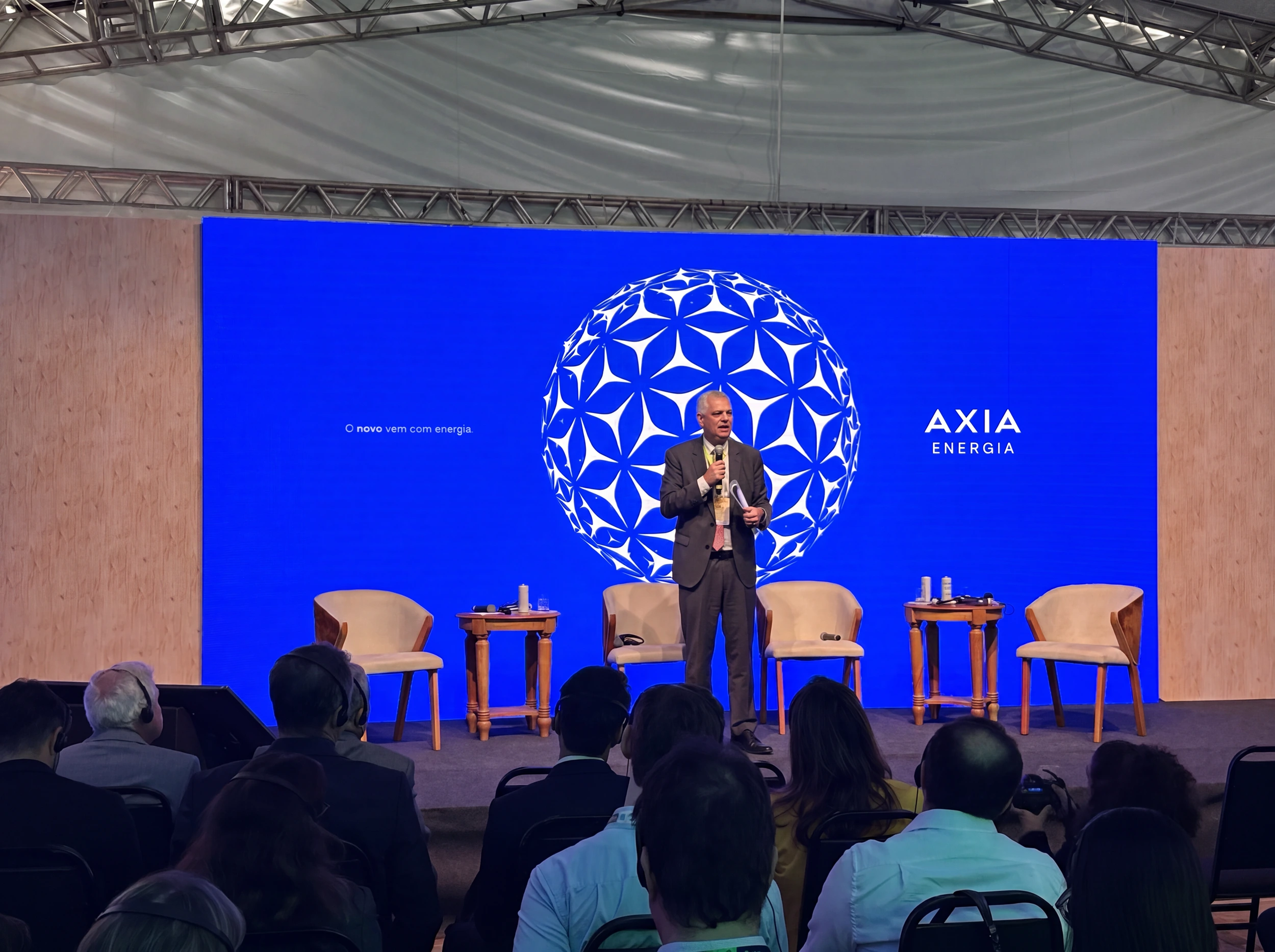 Pessoa palestrando em palco no Energy Summit, com público à frente e fundo azul com o logo da AXIA Energia.