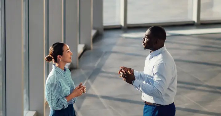 Duas pessoas conversam em um ambiente externo moderno, iluminado pela luz natural, com grandes janelas ao fundo. Uma delas gesticula com as mãos enquanto a outra escuta, em um espaço amplo de circulação.