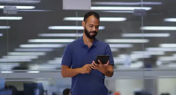 Pessoa em pé em um ambiente de escritório moderno, segura um tablet com as duas mãos enquanto interage com a tela. Ao fundo, há mesas de trabalho e iluminação artificial típica de um espaço corporativo.