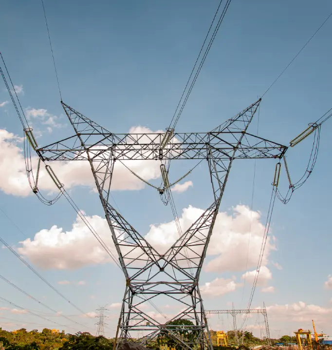 Imagem de uma torre metálica de transmissão de energia elétrica, com vários cabos estendidos, vista de baixo, contra um céu azul com nuvens claras e área verde ao fundo.