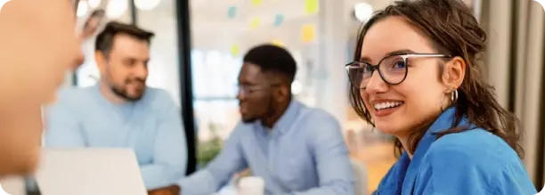 Mulher sorridente usando óculos em primeiro plano, olhando para alguém fora da câmera, enquanto dois colegas conversam ao fundo em um ambiente de escritório moderno.
