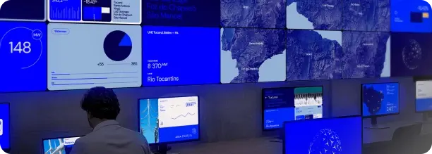 Pessoa de costas observando uma parede com múltiplas telas azuis exibindo gráficos, mapas e dados em um centro de monitoramento.